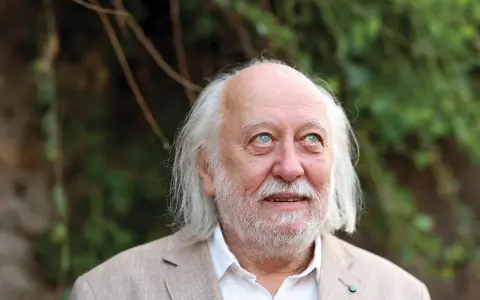 Ritratto di Laszlo Krasznahorkai, scrittore ungherese, con capelli bianchi e barba, sguardo pensieroso rivolto verso l'alto.