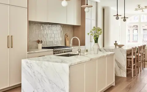 Elegante cocina atemporal con isla de mármol, gabinetes crema y electrodomésticos de acero inoxidable. Un jarrón con flores ilumina el espacio.