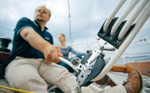 Mann zieht mit kräftiger Hand das Tauwerk am Segel. Ein Mann und eine Frau auf einem Segelboot, die das Tauwerk am Segel bedienen.