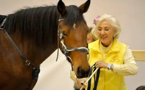 Linda Tellington mit einem braunen Pferd, sie trägt eine gelbe Weste und eine Blume im Haar.
