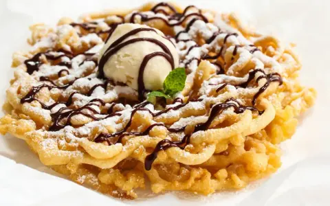 Pyszny funnel cake posypany cukrem pudrem, polany czekoladą i udekorowany gałką lodów waniliowych.