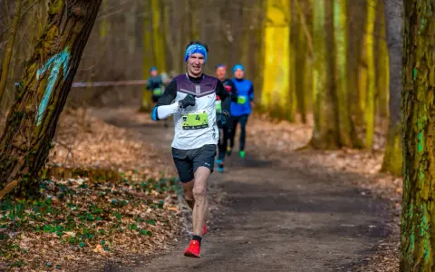Uśmiechnięty biegacz z numerem 4 na koszulce, w niebieskiej opasce, biegnie leśną ścieżką. To bart przedwojewski, który pokonuje trasę biegu.