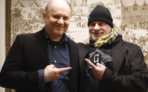 Deux hommes souriants, l'un avec une chemise à pois, l'autre avec un bonnet, se montrent du doigt. Un chanteur de confiance.
