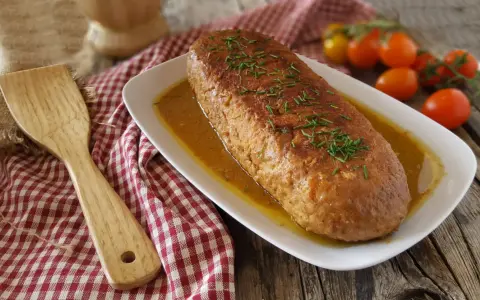 Polpettone in padella dorato, guarnito con erba cipollina, servito su un piatto bianco con salsa. Accanto, una spatola di legno e pomodorini.