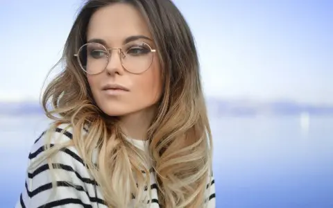 Blondynka w okularach o okrągłych oprawkach, w bluzie w paski, patrzy w bok na tle błękitnego nieba i wody.
