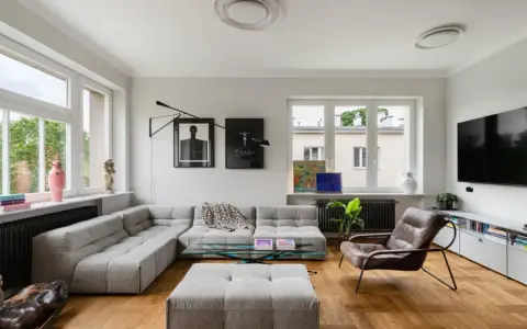 Nowoczesny salon z szarym narożnikiem, szklanym stolikiem i brązowym fotelem. Widać dzieła sztuki, rośliny i telewizor. To przykład udanego **retrofit** mieszkania.