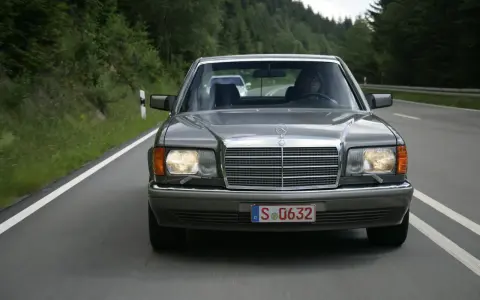 Szary Mercedes W126 jedzie drogą wśród zielonych lasów.