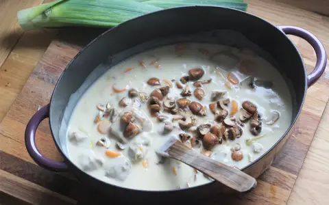 Un plat de blanquette de veau facile mijote dans une cocotte, garni de champignons et de carottes. Un poireau est posé à côté.
