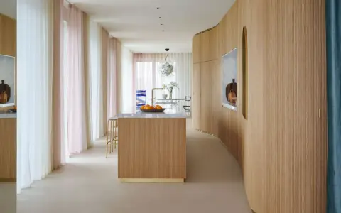 Interno moderno con isola cucina in legno e boiserie curvilinea. Tende leggere color pastello creano un'atmosfera soffusa.