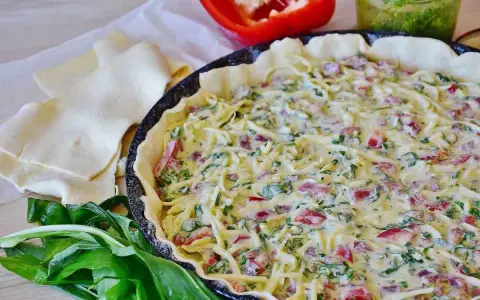 Tarte salée garnie de fromage, poivrons et ail des ours, prête à cuire. Pâte feuilletée et poivron rouge à côté.