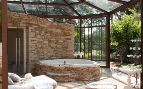 Un spa dans une cave, avec un jacuzzi en pierre, une banquette et une verrière donnant sur un jardin luxuriant.