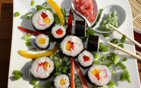 Kolorowe rolki sushi z surimi, papryką i mango, ozdobione szczypiorkiem i papryczkami chili. Obok wasabi i marynowany imbir.