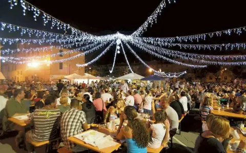 Festa notturna con migliaia di persone sedute ai tavoli, illuminate da festose luminarie. Si celebra la sagra te lu purpu, un evento conviviale.