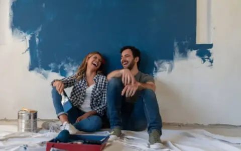 Pareja riendo sentada en el suelo frente a una pared recién pintada de azul. ¡Qué divertido es pintar una habitación!