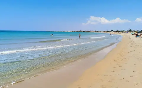 A wide, sandy beach with clear turquoise water and a few people swimming and sunbathing under a bright blue sky.