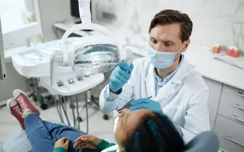 Dentysta w masce i rękawiczkach bada pacjenta w gabinecie stomatologicznym.
