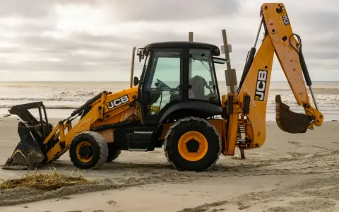 Żółta koparko-ładowarka firmy JCB stoi na piaszczystej plaży, gotowa do pracy.
