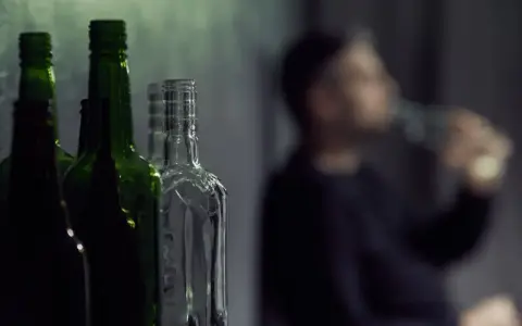Puste butelki na pierwszym planie, w tle niewyraźna postać pijąca alkohol. Gdzie jest bezpieczna ilość alkoholu?