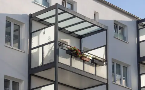 Nowoczesny balkon dostawny z przeszklonym zadaszeniem i balustradą, ozdobiony skrzynkami z kwiatami. Sprawdź balkon dostawny cena.
