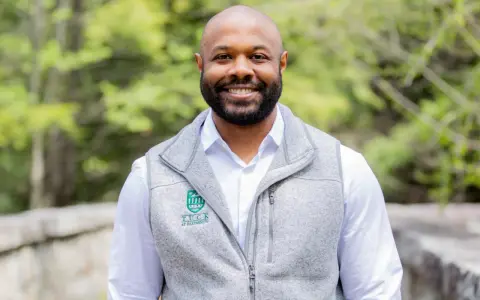 A smiling man with a beard wears a grey vest over a white shirt. The vest has a Tuck at Dartmouth logo, suggesting a curated professional image.