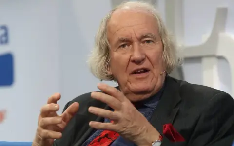 Dieter Borchmeyer, mit seinem charakteristischen langen Haar und einem gestreiften Anzug, gestikuliert lebhaft, während er spricht.