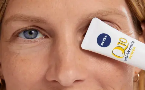 Un tube de crème pour les yeux Nivea Q10 Anti-Wrinkle Power est tenu devant l'œil d'une femme, masquant les rides pattes d'oie.