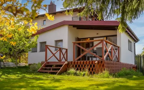 Bungalow z drewnianym tarasem i schodkami, otoczony zielonym ogrodem i kwitnącym drzewem.