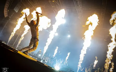 Un chanteur de groupe hard rock des années 2000, micro en main, saute sur scène entouré de jets de flammes spectaculaires.