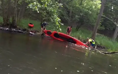 Ludzie wyciągają czerwoną kajak z wody. W tle widać drzewa i drugą czerwoną łódkę. Woda jest ciemna, jakby to było jezioro smołowe.