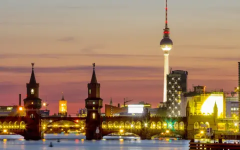Berlin Kreuzberg bei Sonnenuntergang: Oberbaumbrücke, Fernsehturm und beleuchtete Gebäude spiegeln sich im Wasser.