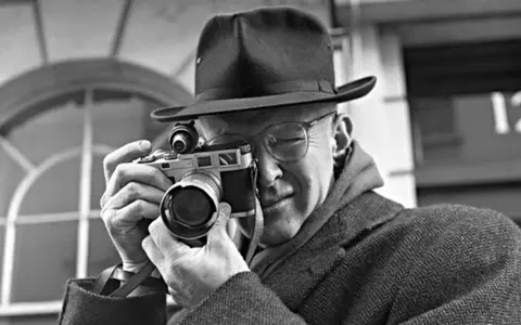 Mężczyzna w kapeluszu i okularach trzyma aparat, gotowy do zrobienia zdjęcia. Wśród znanych fotografów, jego postawa sugeruje skupienie i profesjonalizm.