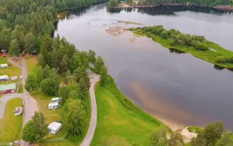 Vacker vy över Selholmens camping med husbilar och stugor vid en sjö omgiven av skog.