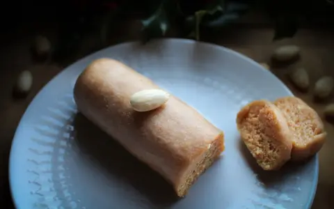 Un bâtonnet de pâte d'amande, garni d'une amande entière, repose sur une assiette blanche. À côté, deux tranches de ce délice, parfait pour un fond de tarte amande.