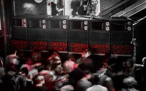 Altavoces apilados con luces rojas "GREEN LIGHT" y gente borrosa bailando. Un sistema de sonido que parece dar problemas.