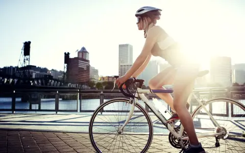 A female cyclist enjoys a sunny ride along a waterfront path with a city skyline in the background.