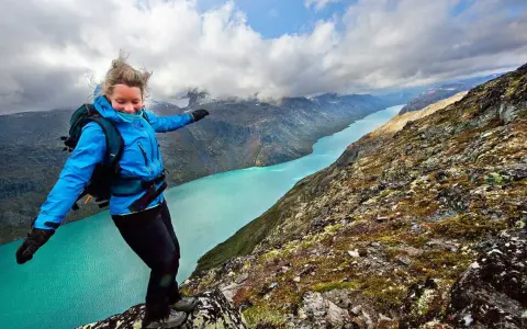 En kvinna med ryggsäck balanserar på en klippkant med en turkos sjö och berg i bakgrunden, en vy som påminner om Besseggen i Norge.