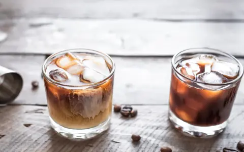 Dwa drinki na drewnianym stole: jeden to kremowy White Russian, drugi to ciemny Black Russian.