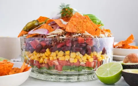 Schichtweise aufgebauter Taco Salat im Glas mit Hackfleisch, Bohnen, Mais, Tomaten, Salat, Nachos, Jalapeños und Avocado.