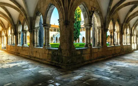 Gotische Arkaden im Kloster Batalha, Portugal. Sonnenschein fällt auf den Steinboden und beleuchtet den grünen Innenhof.