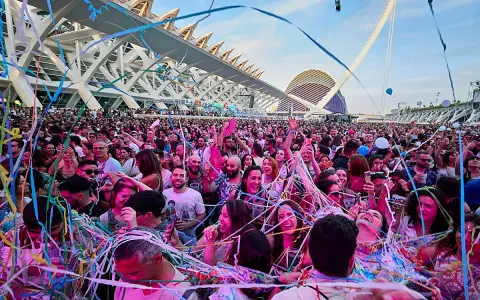 Multitud disfrutando de un fan music fest bajo confeti y luces, con arquitectura moderna de fondo.