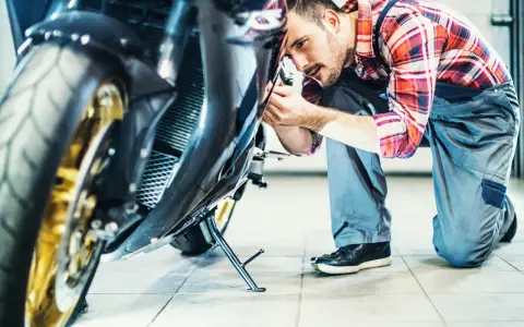 Mechanik w kratkę wykonuje przegląd motocykla, pochylając się nad jego przednią częścią.