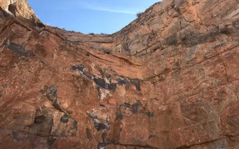 Acantilado de roca rojiza con vetas oscuras, mostrando diferentes tipos de canteras y la estratificación geológica bajo un cielo azul.