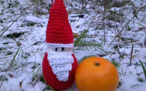 En virkad tomteluva med vitt skägg står i snön bredvid en apelsin.