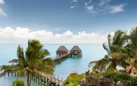 Dwa domki na wodzie połączone pomostem, otoczone palmami. Spokojne morze i błękitne niebo z chmurami. Zanzibar, pora deszczowa, ale pogoda dopisuje.