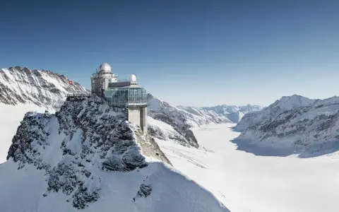 A modern building with glass walls and domes sits atop a snowy mountain, offering breathtaking views of the surrounding peaks and a vast glacier. This is the top of Europe, a place of incredible altitude.