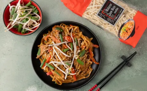 Pyszny makaron chow mein z warzywami i kiełkami fasoli na czarnym talerzu, obok paczka makaronu i miseczka kiełków.