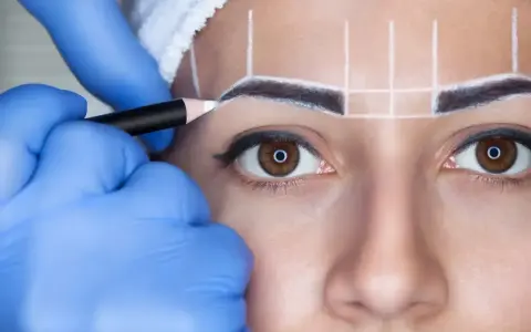 Stylist w niebieskich rękawiczkach rysuje kredką idealny kształt brwi przed zabiegiem henny.