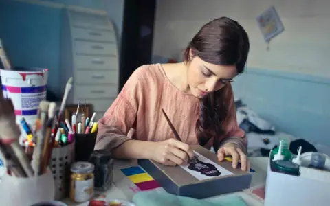 Une jeune femme se déclare artiste libre, concentrée sur sa toile, entourée de pinceaux et de peintures.