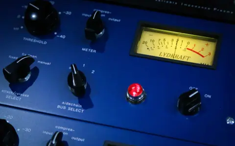 Gros plan sur un compresseur audio bleu avec des boutons, un VU-mètre et une lumière rouge allumée.