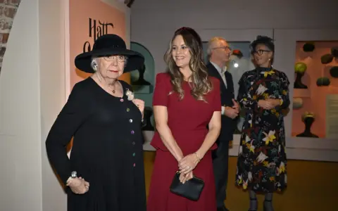 Prinsessan Sofia beundrar en utställning med hattar, där en äldre dam bär en elegant svart hatt.